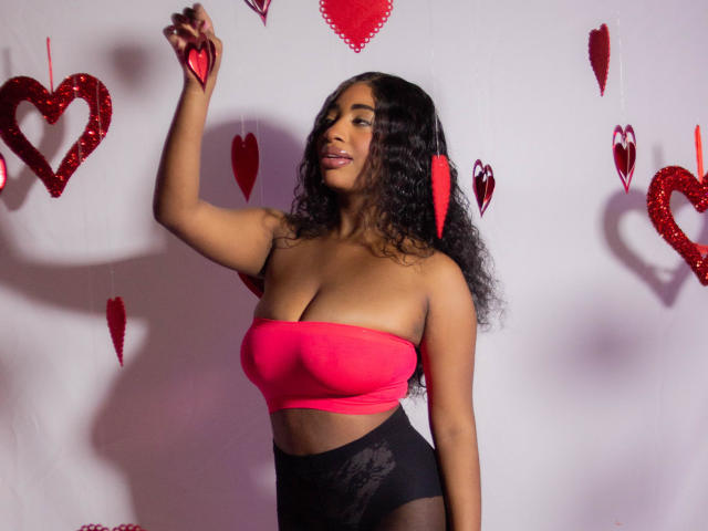 Charlote Brown, con i capelli neri ricci, indossa un top rosa senza maniche, mostrando il suo seno prosperoso e biancheria intima di pizzo nera, tiene un cuore rosso, sorride e alza un braccio..