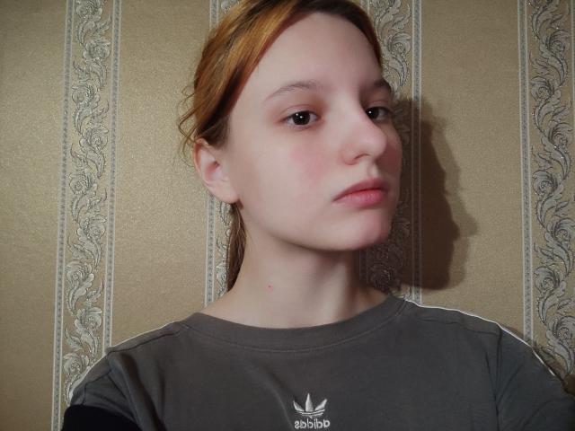 KodikMya's face with light skin, reddish-brown hair, and a slight frown. She's wearing a gray Adidas shirt with a white logo. Her shadow is on the wall.