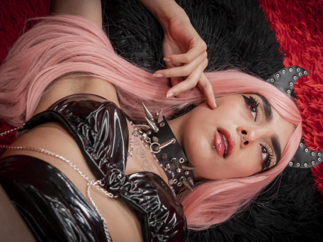 Ariana Spencer, con i capelli rosa, una giacca di vinile nero, un collare con borchie e una catena, sdraiata su pelliccia nera e rossa, che tocca il naso, con un anello al naso e labbra rosse lucide..