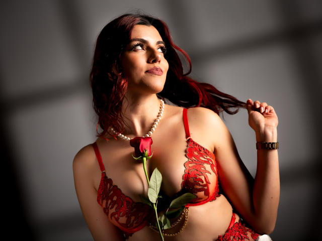 ZoeeMar with red lace bra, pearls, and a rose between her tits, holding her dark red hair, looking up with a soft smile. She's sexy and confident.