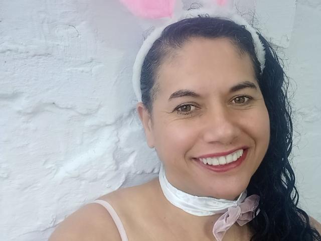 AmieLove with dark curly hair, white headband, pink bunny ears, white choker with pink bow, smiling, pink lipstick, white textured wall in background.