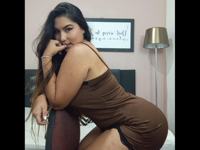 Maria Rowe, met lang donker haar, in een bruine jurk, die haar lip aanraakt, laat haar achterwerk zien, zwarte kanten lingerie, en haar ronde figuur, leunend op een stoel, slaapkamer, sexy..