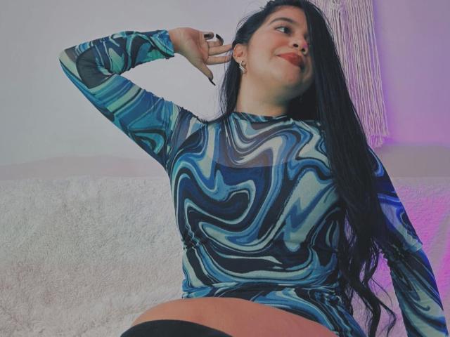 SweetYolandaX with long black hair, wearing a blue, black, and white marbled long-sleeve top, making a peace sign, smiling, sitting on a white couch, showing her ass.