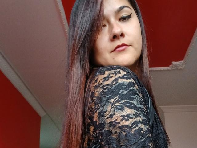 FreeMystic69 com cabelos castanhos longos, vestindo uma blusa preta de renda, olhando por cima do ombro, com delineador, teto vermelho, moldura branca, sorrindo levemente, mostrando o nariz e a parte superior do peito..