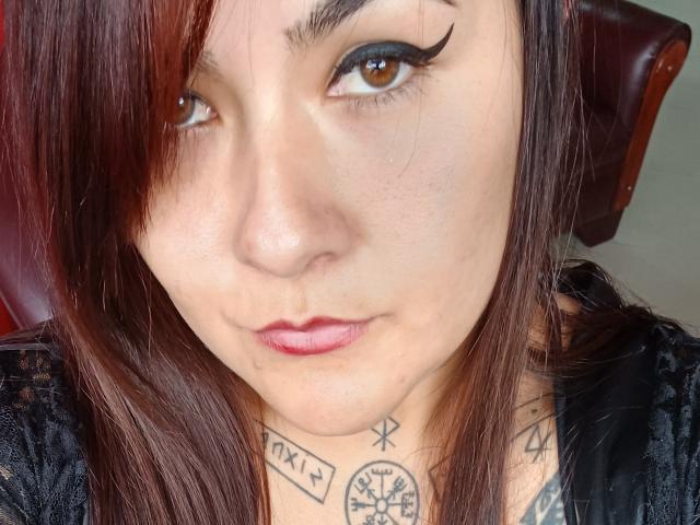 FreeMystic69 heeft lichtbruine ogen, rode lippenstift en donkere eyeliner. Ze heeft tatoeages op haar nek en borst, en bruin haar met rode highlights, en draagt een zwarte top..
