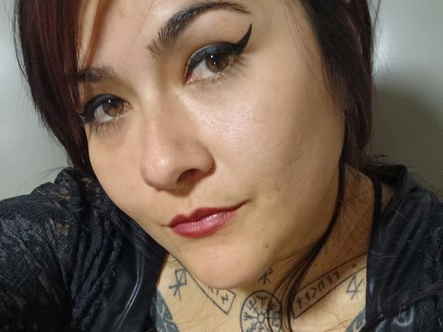 FreeMystic69 has dark eyeliner, red lipstick, and tattoos on her chest. She has light brown skin, brown eyes, and red-brown hair. She's wearing a black lace shirt.