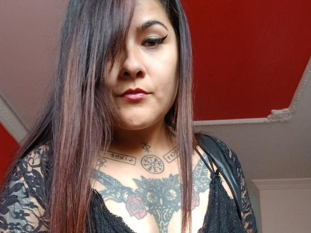 FreeMystic69 a de longs cheveux bruns, porte un haut en dentelle noire, a des tatouages sur la poitrine et a un air sérieux. Ses seins sont visibles, et le tatouage sur sa poitrine comprend une horloge et une croix. Le plafond au-dessus est rouge..