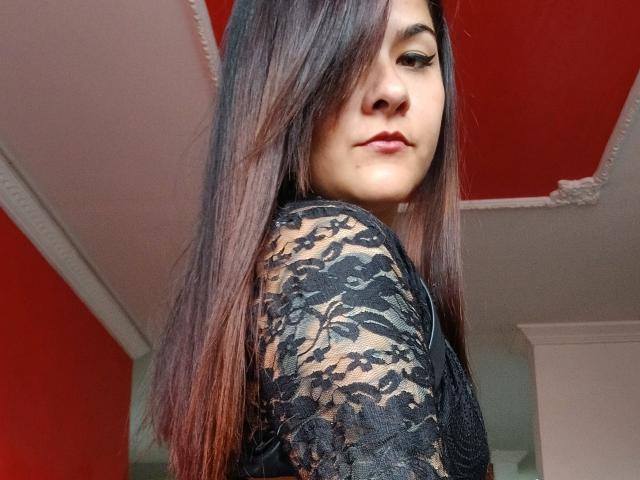 FreeMystic69 heeft lang bruin haar, draagt een zwarte top van kant, kijkt over haar schouder met een lichte grijns, en heeft een rode plafond, witte kroonlijsten en subtiele make-up..
