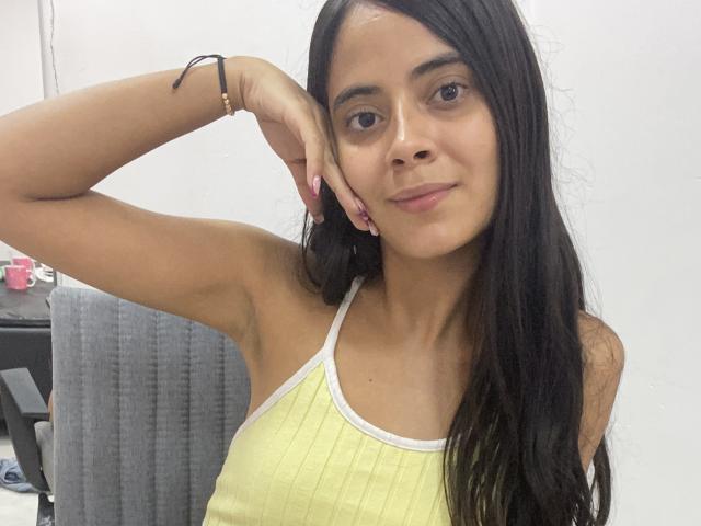 TamaraPeach, avec de longs cheveux noirs, une peau claire, de petites seins, portant un débardeur jaune côtelé, un léger sourire, le bras droit levé, un bracelet noir, du vernis à ongles rose, dans un cadre décontracté..