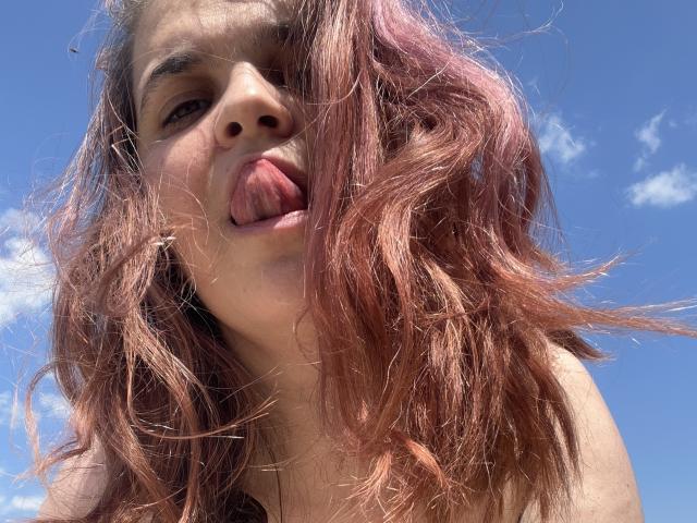 NaturalTessa with pink, wavy hair, sticking out her tongue, has a light skin tone, and is looking down at the camera, showing her nose and part of her upper chest.