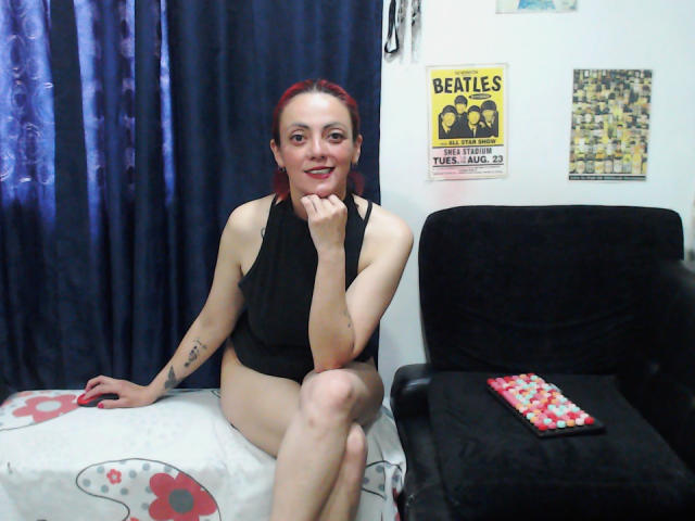 SamanthaSwan with red hair, black sleeveless top, and black panties, sitting on a bed with white and red floral blanket, black armchair, and red and white candies.