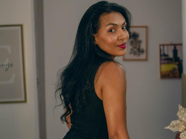 AlysonStone with long black hair, wearing a black sleeveless top, turning to look over her shoulder with red lipstick, medium tits, and a slight smile in a room.