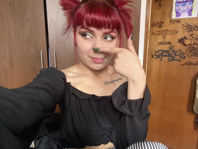 KallySpice with red pigtails, black off-shoulder top, nose ring, tattooed chest, making a peace sign, smiling, visible breasts, black and white striped chair in foreground.