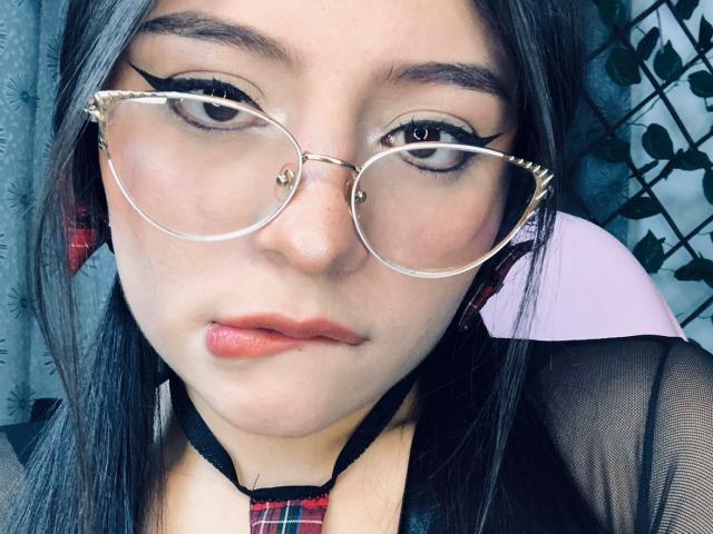 ElizabethCooper with pale skin, black hair, and red lips, wearing gold-rimmed glasses, a black mesh top, and a red plaid choker, looking serious with slightly parted lips.