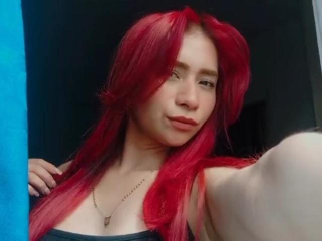 MelannyTaylorr with red hair, light skin, small tits, wearing a black top, necklace, smiling slightly, taking a selfie, blue curtain on the left, indoors, soft lighting.