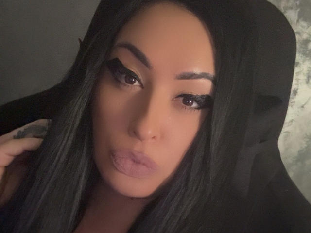 DevilleDawn with long black hair, thick eyebrows, and full lips wearing dramatic eyeliner, looking down with a pouty expression, her nose slightly centered, in a dimly lit photo.