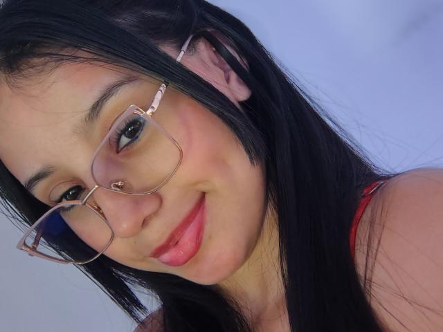 LilyC has black hair, glasses with pink frames, a nose ring, red lipstick, and a cute smile. Her tits are not visible, but she's wearing a red top.