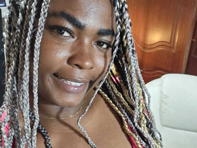 KarlaFlorez with dark skin, long blonde and brown braids, smiling, glossy lips, small gold necklace, visible teeth, black eyebrows, looking at camera. Boobs partially visible, pink top.