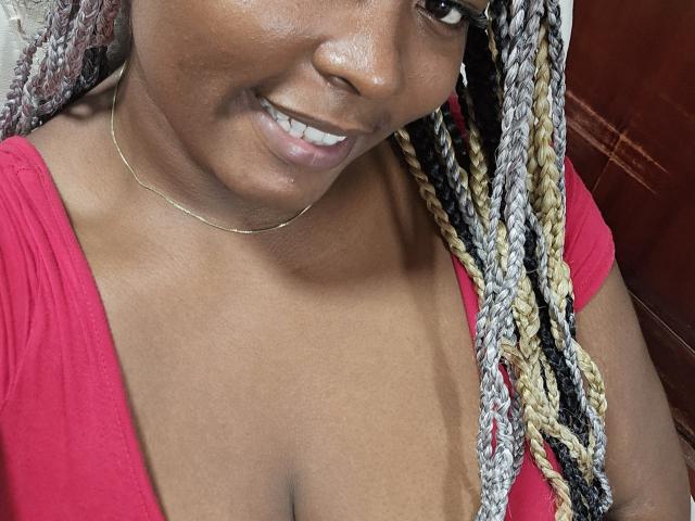 KarlaFlorez with dark brown skin, smiling, wearing red top, showing cleavage, and blonde black gray braids, wearing thin gold necklace, has shiny skin and white teeth.