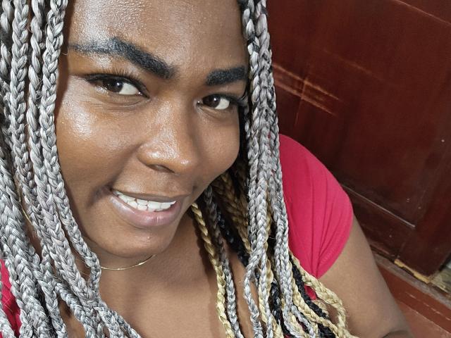 KarlaFlorez has dark brown skin, silver braids, thick eyebrows, and a smiling face with white teeth, wearing a red shirt. Her chin has a small, visible bruise.