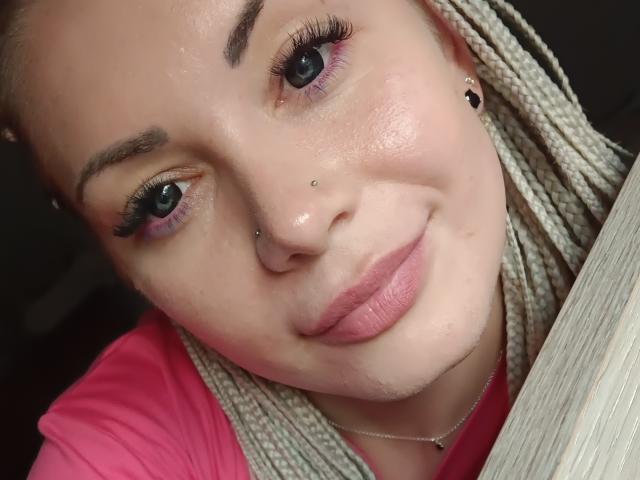 BlossomPie has light skin, braided blonde hair, purple eyeshadow, black eyeliner, long eyelashes, nose piercing, lip piercing, pink shirt, and a silver necklace, smiling softly.