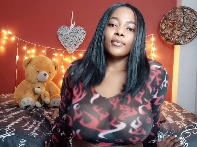 MacarenaBoobs with black straight hair, wearing a black and red flame-patterned top, sitting on a bed with a brown teddy bear, warm string lights, and a heart decoration.