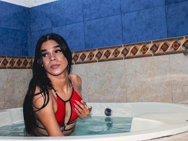 KataHot69 with long black hair, red strappy bikini top, wet skin, and small tits, sitting in a white bathtub with water, looking up with a slight smile.