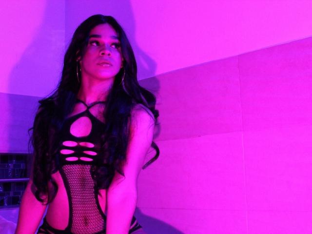 KataHot69 with long black hair, wearing a black mesh lingerie top with cutouts, standing under pink and purple lighting. Her ass and breasts are visible through the mesh.