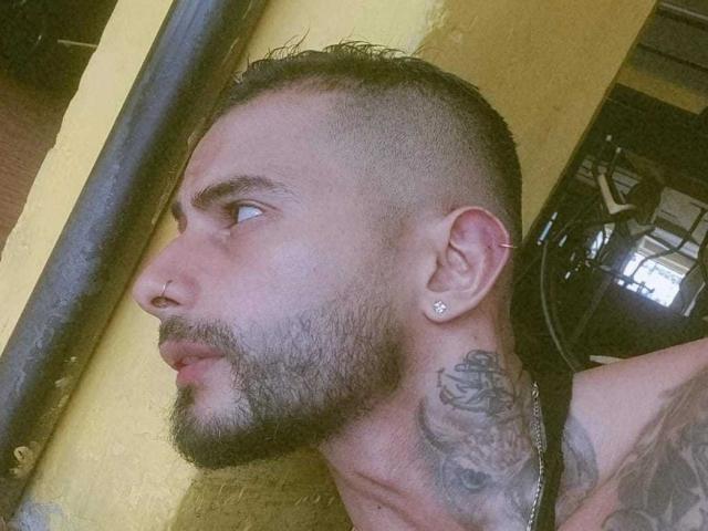 MaxJhonson's side profile with a shaved head, beard, nose ring, ear piercing, tattoo on the neck, and a necklace, looking to the left in a dimly lit room.