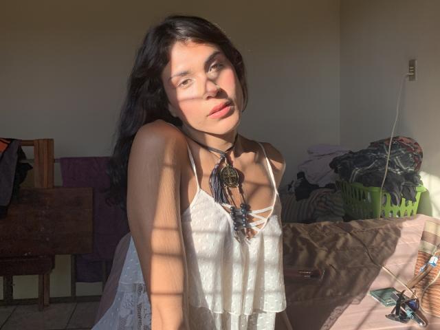 VensFun, avec de longs cheveux noirs, baignée de soleil, portant un haut en dentelle blanche, un collier de perles, le visage dans l'ombre, et de douces lèvres roses, assise dans un rayon de soleil..