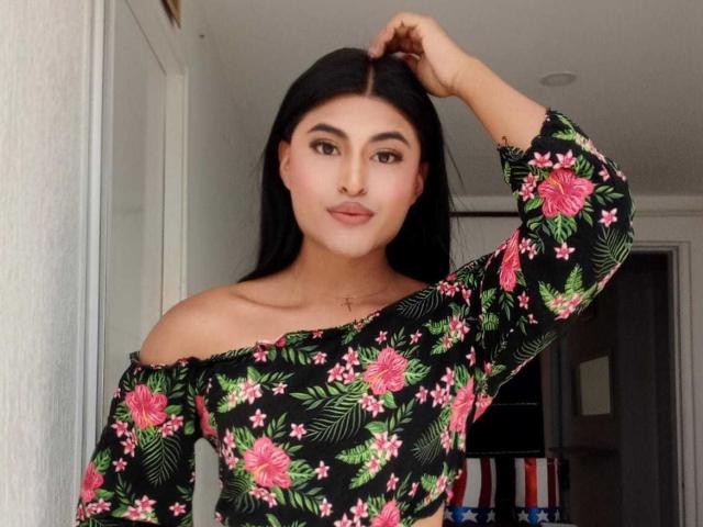 MichelleDenise with long black hair, brown eyes, and pink floral off-shoulder top, hand in hair, standing, looking at camera, subtle smile, cute, sexy.