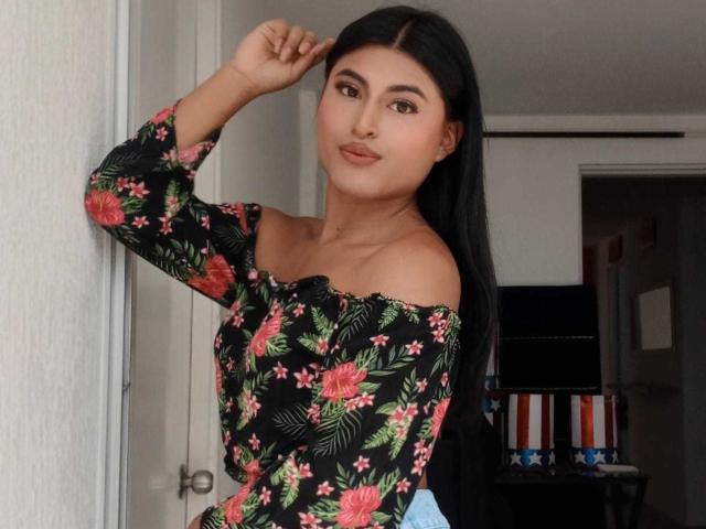 MichelleDenise with long black hair, wearing an off-shoulder floral top, has small tits and a cute face, standing in a doorway, touching her hair with a subtle smile.