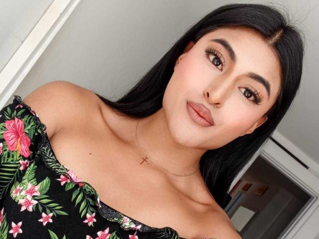 MichelleDenise with long black hair, brown eyes, and full lips, wearing a floral off-shoulder top, gold necklace, and subtle makeup, smiling at the camera, showing her tits.