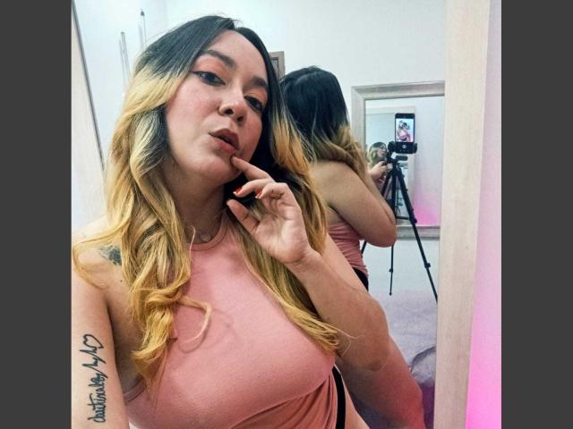 KimikoMoon with blonde ombre hair, pink top, finger on lips, tattoo on arm, mirror selfie, camera in background, sexy, tits visible, reflection shows camera and tripod.
