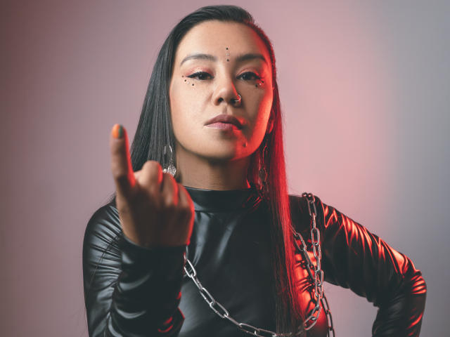 CharlotteJoliie with straight black hair, black leather outfit, chain necklace, nose ring, and freckles, pointing index finger forward, red and gray lighting, serious expression.