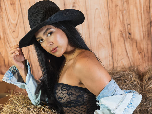 SamanthaEva69 with long black hair, wearing a black lace strapless top, denim jacket off-shoulder, and black cowboy hat, sitting on hay, looking at camera, showing her tits.