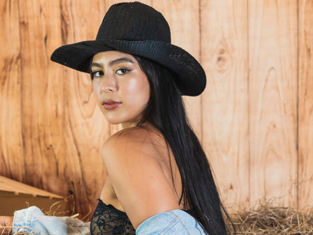 SamanthaEva69 with long black hair, wearing a black hat, lace top, and blue jacket, showing off her bare shoulders and looking over her shoulder with a pouty expression.