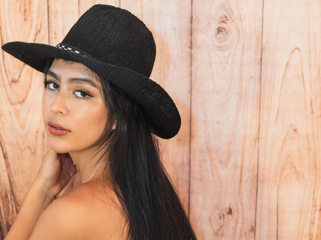 SamanthaEva69 with long black hair, wearing a black woven hat, looking over shoulder, brown eyes, subtle makeup, tan skin, hands near neck, wooden background, no clothes visible.