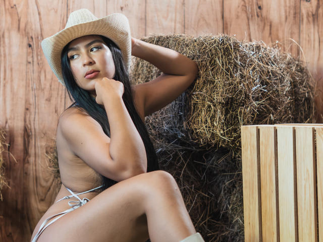 SamanthaEva69 with long black hair, tan skin, wearing a straw hat, sitting topless, showing her small tits, with a hand on her chin, next to hay and wood.