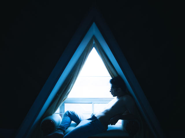 TonyDan con seno e glutei, seduto in un telaio di finestra a forma di triangolo, che indossa una canotta, jeans blu e occhiali, illuminato da una luce blu soffusa, in una stanza buia..