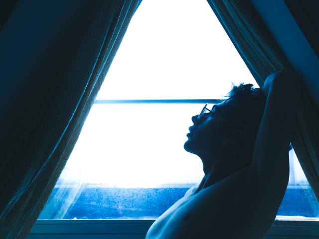 Tonys Profil, ohne Hemd, in blauem Licht, mit erhobenen Armen, zeigt eine glatte, trainierte Brust und das Seitenprofil des Gesichts. Im Schatten vor einem hellen Fenster, umrahmt von grünen Vorhängen..