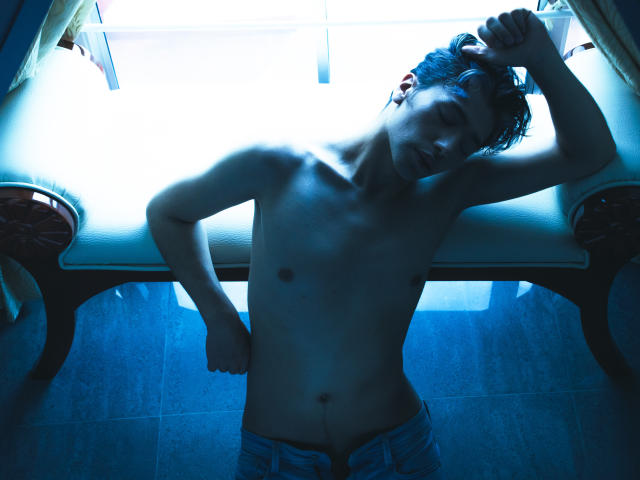 TonyDan é um homem sem camisa, com corpo magro, em pé sob uma luz azulada, vestindo calças jeans abertas, com a mão no cabelo, com uma expressão cansada, e apoiado em um banco branco..