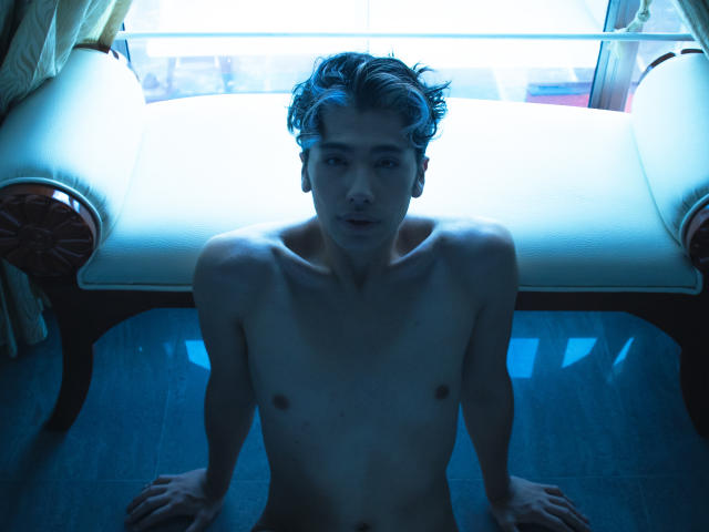 TonyDan is a skinny, naked dude with small tits and no ass, sitting on the floor with blue lighting, looking at the camera. He has short, wavy hair.