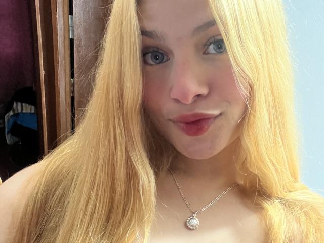 ShantalSummer has blonde hair, blue eyes, and pink lips with a slight smirk. She's wearing a silver necklace with a gem. Her tits are not visible.