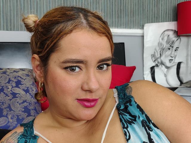 CathaLoreth with brown hair in a bun, pink lipstick, green lace top, tattoos, earrings, brown eyes, slight smile, red pillow, black-and-white Marilyn portrait, gray wall.