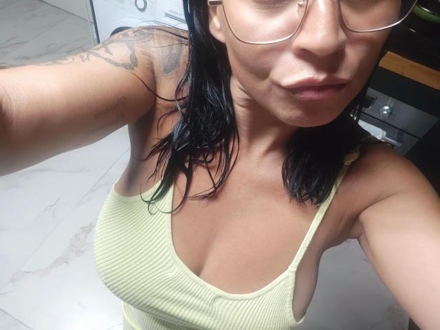 CleopatraUnica-hot with tan skin, black hair, glasses, and a yellow ribbed tank top showing her tits. Tattoo on left arm. Marble floor. Pouty lips. Selfie.