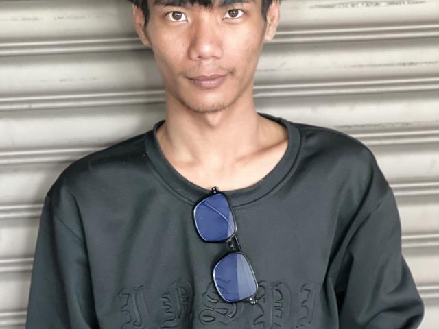 BiMonsterCock with medium brown skin, short black hair, and slight stubble, wearing a black shirt with blue aviator sunglasses hanging from his neck, smiling subtly.