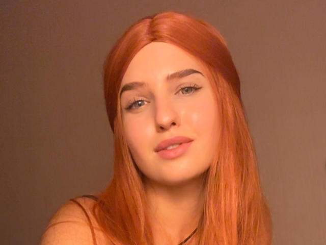 RosyAlma, con cabello rojo, piel clara y labios rosados y gruesos, mostrando una sonrisa sutil, con pestañas largas y un pequeño collar negro sobre un fondo marrón..