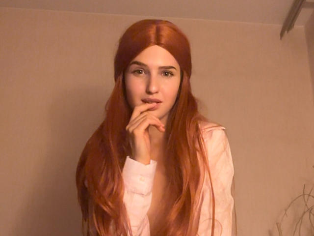 RosyAlma with long red hair, wearing a pink shirt, has her hand near her mouth, looking slightly bored or thoughtful. Her tits are small, she's cute, and has light skin.