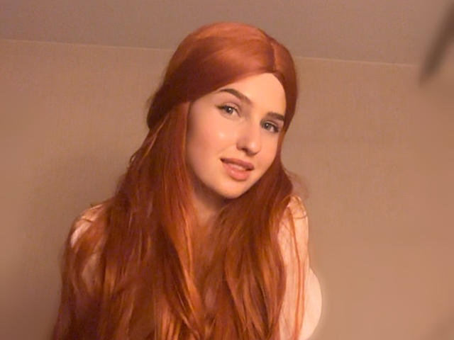 RosyAlma tiene el cabello largo y rojo, piel clara y una sonrisa encantadora. Está mirando a la cámara con una ligera inclinación de la cabeza. Lleva una blusa blanca sin mangas..